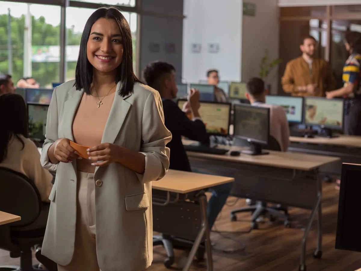 Colaboradora sonriente en una oficina abierta, con compañeros trabajando en sus computadoras